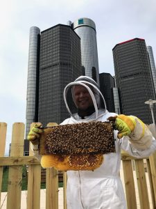Brian Peterson-Roest en un traje completo de abeja sostiene un marco de abejas melíferas frente a un edificio alto en el fondo