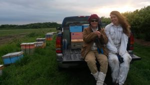 Julia Common con sombrero rojo y Sarah Common en traje de abeja se sientan en la parte trasera de una camioneta en un campo con colmenas a su izquierda
