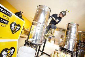 Kristy Lynn Allen in black helmet with bee tentacles & yellow & black striped socks pedals a metal container outputting honey
