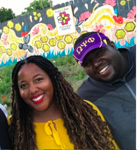 Nicole Lindsey en amarillo y Timothy Jackson en una visera púrpura y amarilla sonríen frente de un mural de abeja pintado en una cerca
