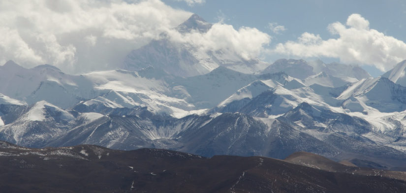 This photo shows Mount Everest as seen from a distance. It is a large, pyramid-shaped, craggy peak with many smaller snow-covered peaks in the foreground. The peak of Mount Everest is partially occluded by clouds.