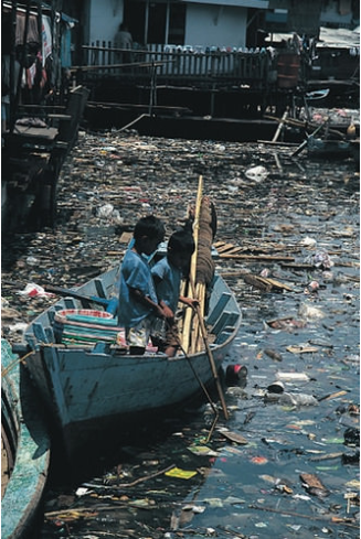 Two boys are in a small boat that floats on a river with trash floating on the surface.