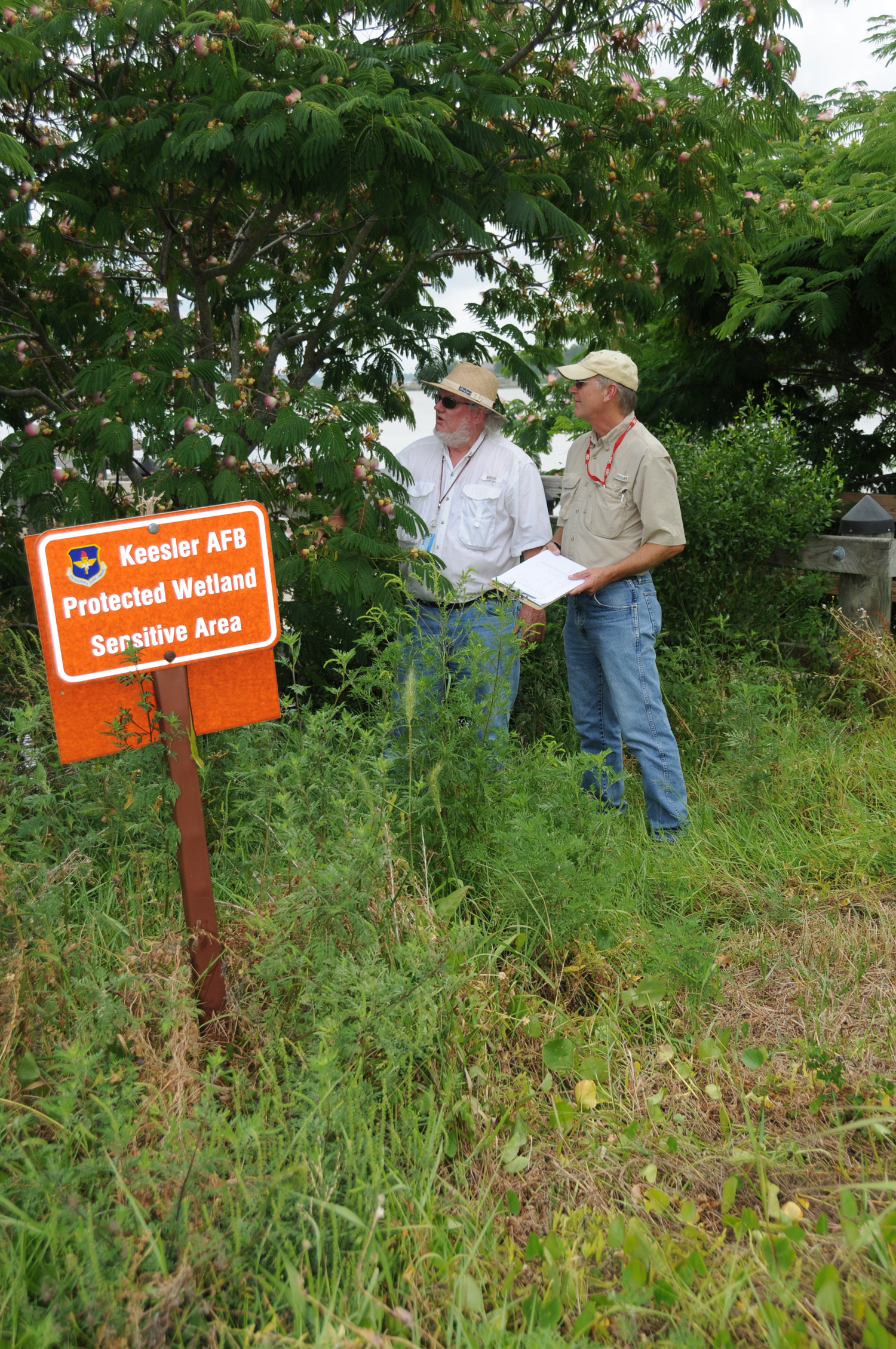 Two men stand in dense vegetation underneath trees. A sign reads, "Keesler AFB, Protected Wetland, Sensitive Area".