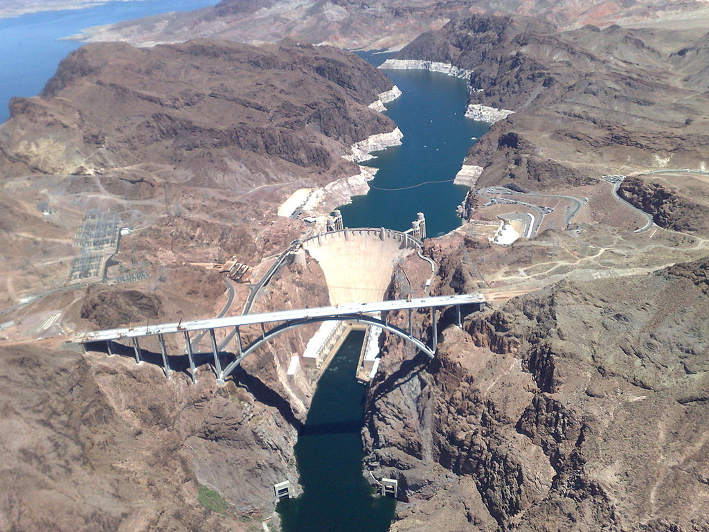 Aerial view of the hoover dam shows a concrete structure obstructing a wide channel of water (the Colorado River). Lake Mead is in the upper right corner.