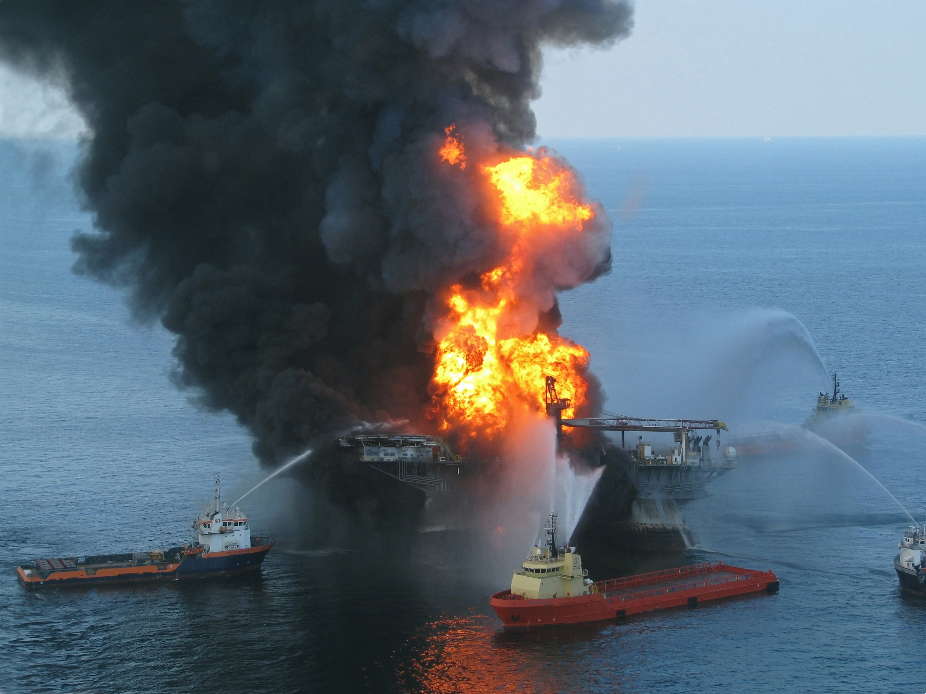 Ships spray a fire on the ocean resulting from an oil spill. A dark plume of smoke rises from the fire.