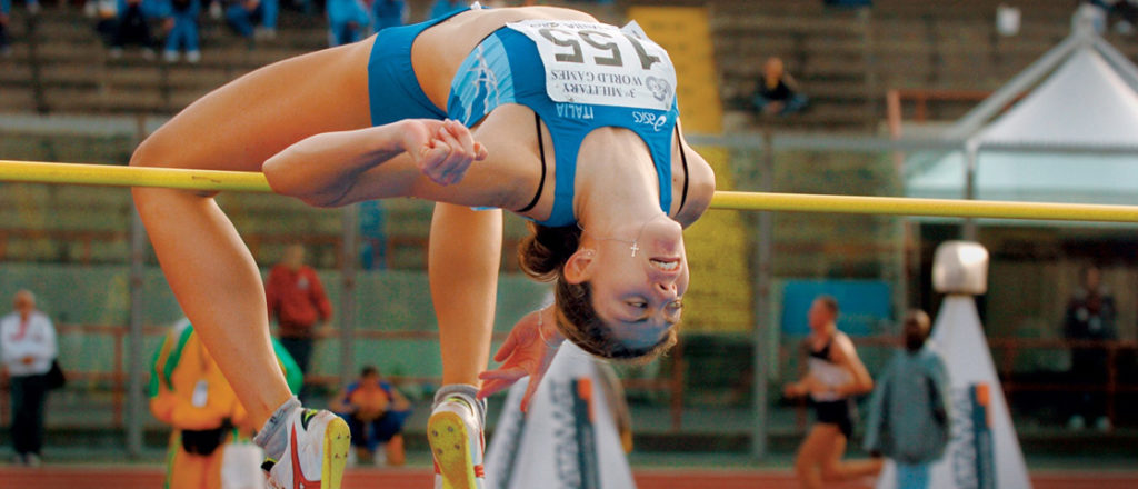 Illustration shows a woman, upside-down with an arched back, going over a pole vault.