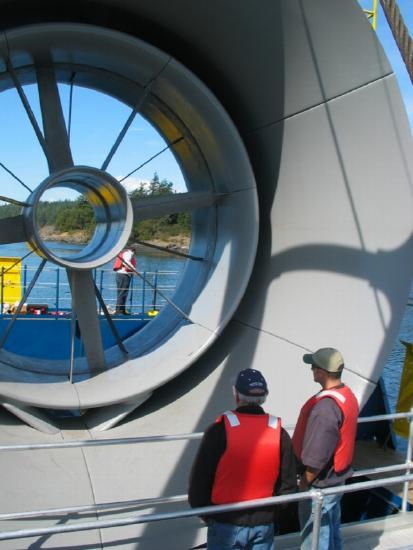 Two men in life vests stand in front of a giant turbine. The ocean is visible behind it.