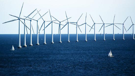 A row of tall, white wind turbine, each with three blades, floating at sea. Sailboats are tiny in comparison.