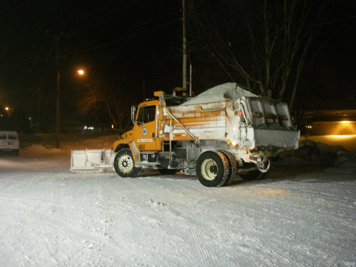 A snow road and large truck with a snow plow in the front and a bed full of salt