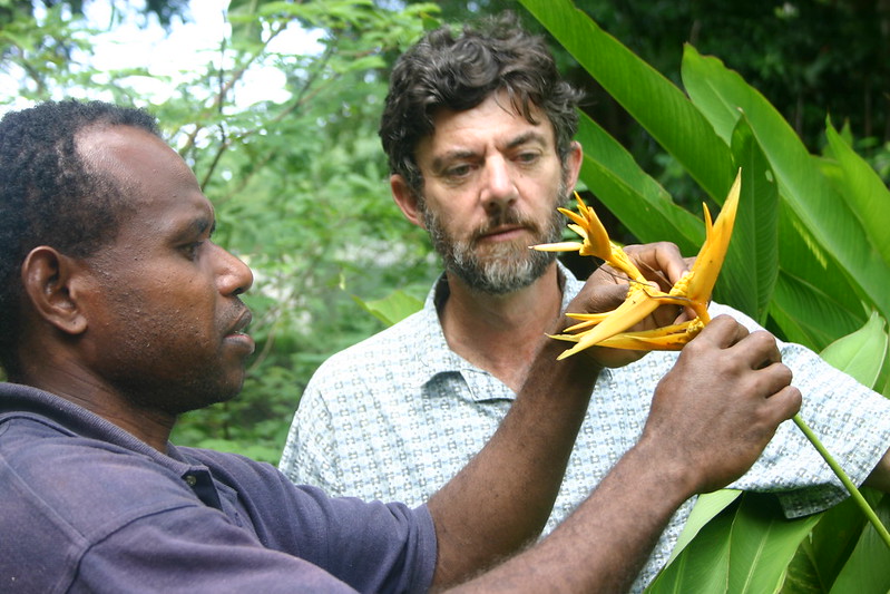 A horticulturalist holds a tall, orange flower while speaking to a volunteer advisor