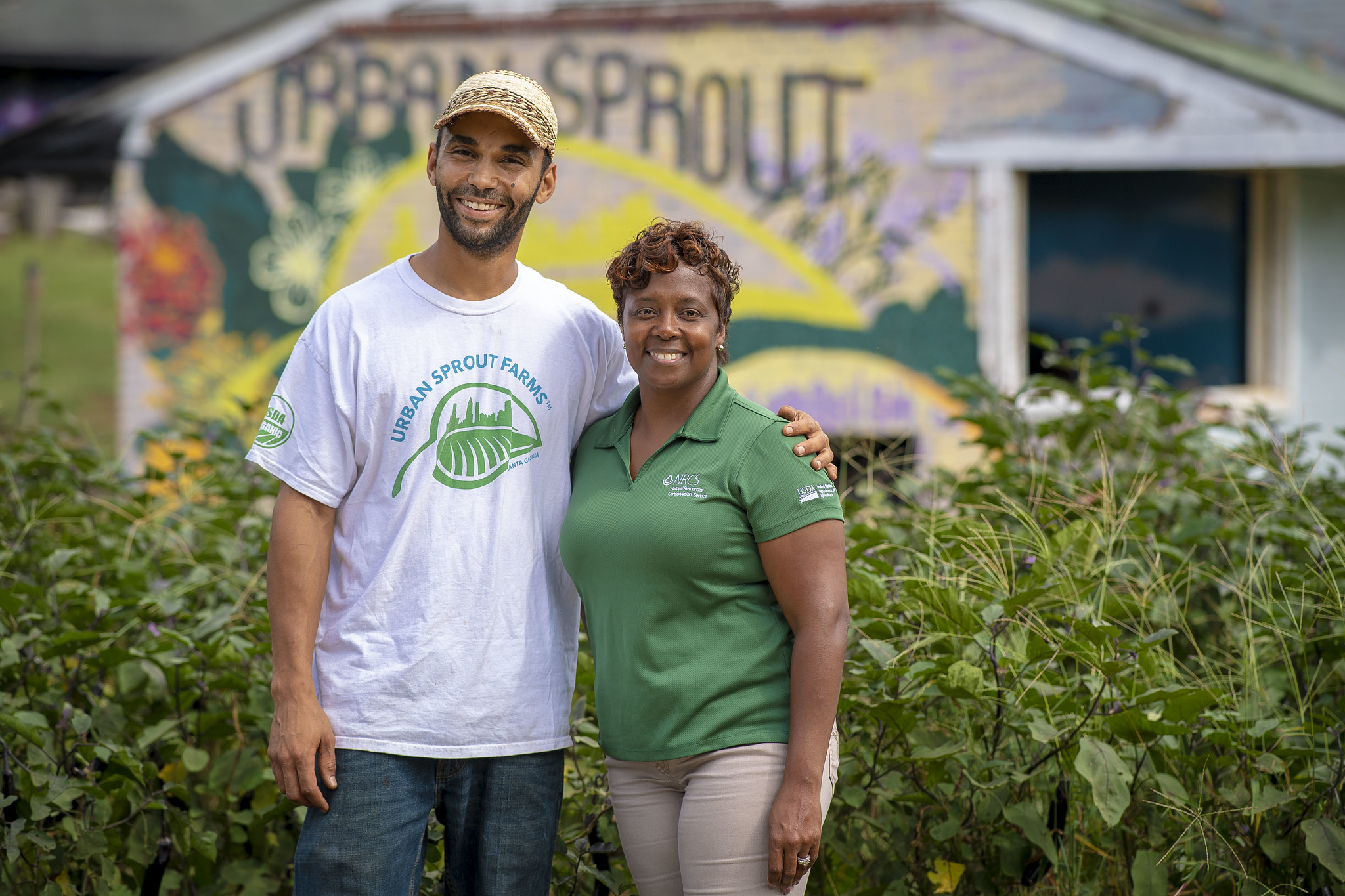A USDA employee and organic farmer with a shirt that says "Urban Sprout Farms" stands in front of a farm.