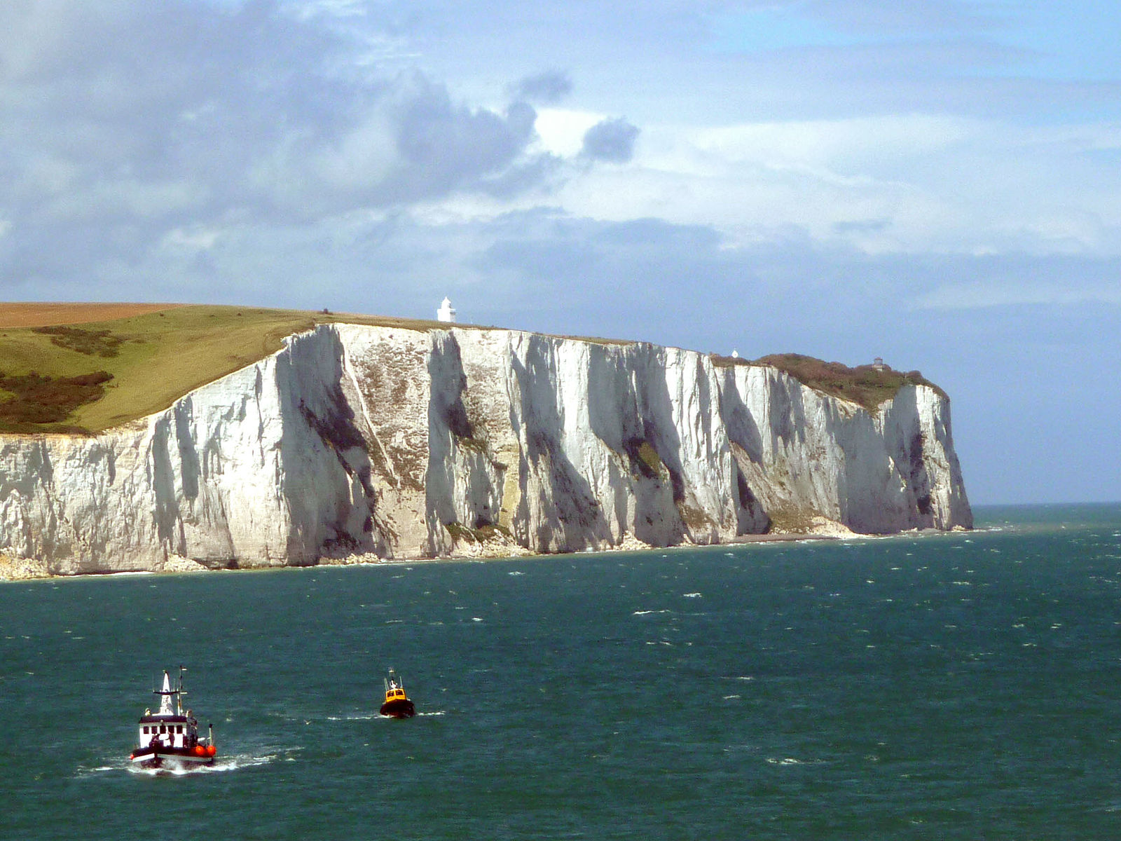 A photo of a steep, white cliff with grass growing on the hillside, below the cliff are the blue water and two boats