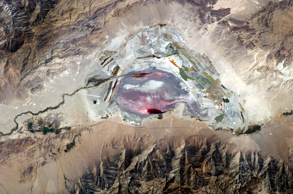This astronaut photograph highlights the mostly dry bed of Owens Lake, located in the Owens River Valley between the Inyo Mountains and the Sierra Nevada. Shallow groundwater, springs, and seeps support minor wetlands and a central brine pool. Two bright red areas along the margins of the brine pool indicate the presence of halophilic (salt-loving) organisms known as archaeans. Gray and white materials within the lake bed are exposed sediments and salt crusts. The nearby towns of Olancha and Lone Pine are marked by the presence of green vegetation, indicating a more constant availability of water.