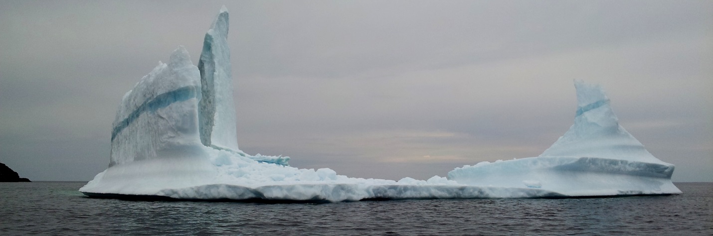 A floating ice burg sits in the center of this photo. The center of the ice burg is thin, with a peak of ice on either side. 