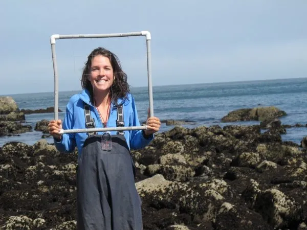 A woman wearing waders is smiling while holding a square of pipes with a rocky coast as her background.
