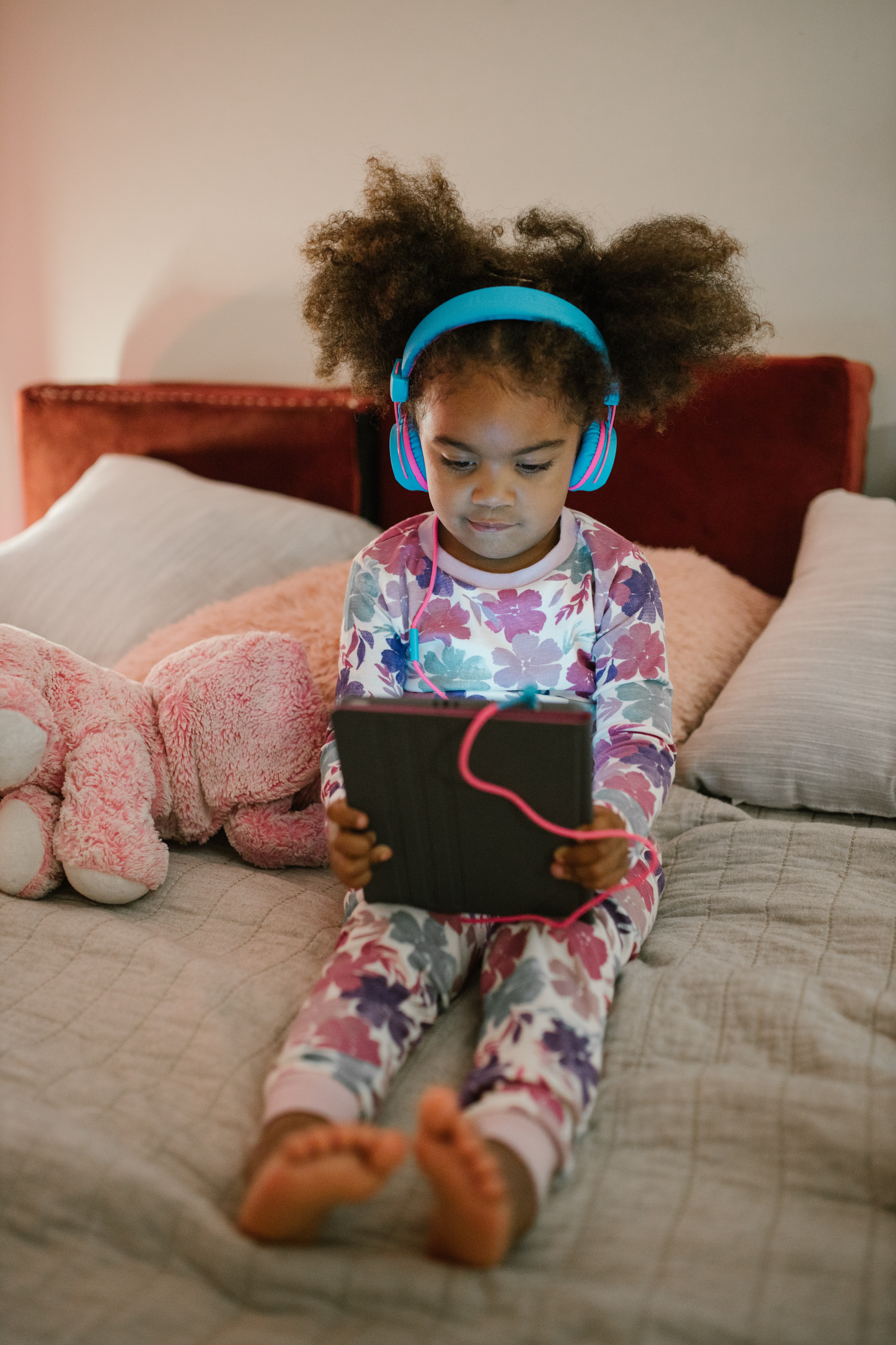 Photo of a little girl with curly hair, sitting on a bed wear pajamas with headphones, looking at a digital tablet.