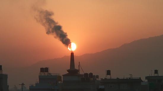 A photograph shows the silhouette of a factory emitting smoke into a cloudy sky as the sun sets.